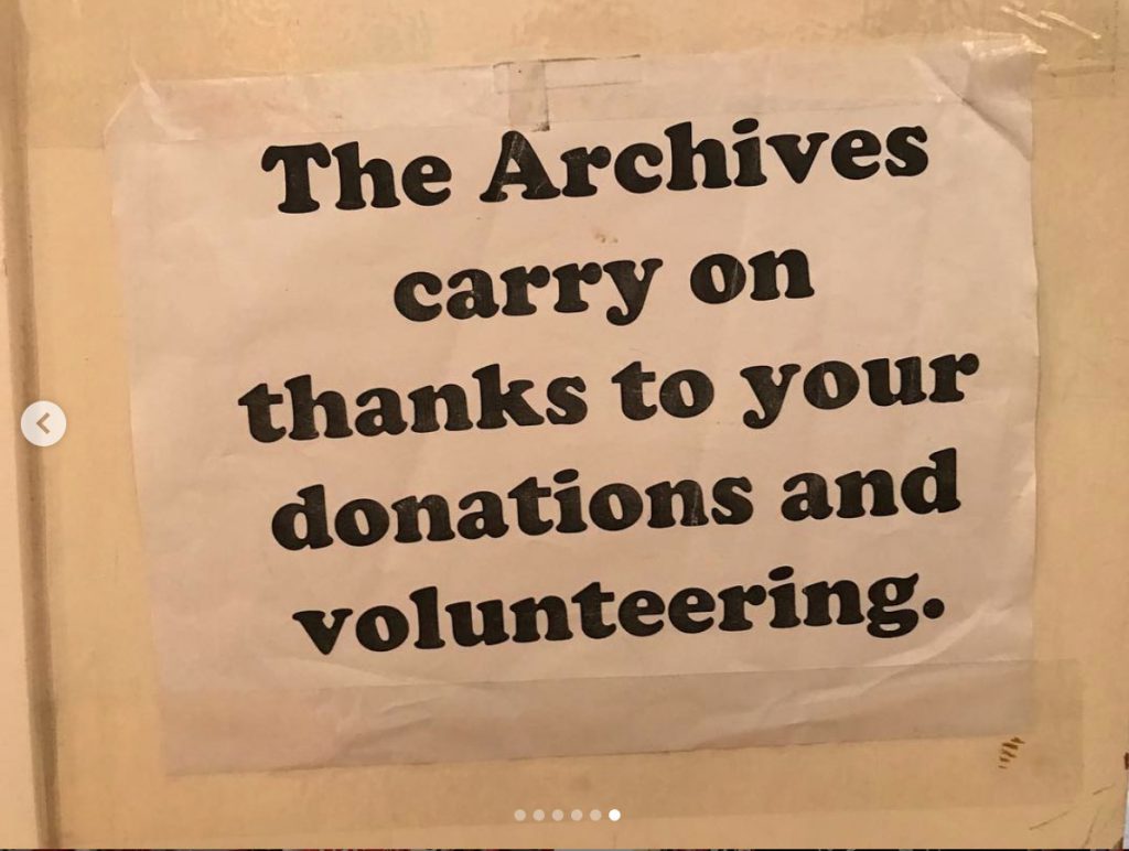 A paper sign taped to a wall that reads "The Archives carry on thanks to your donations and volunteering."