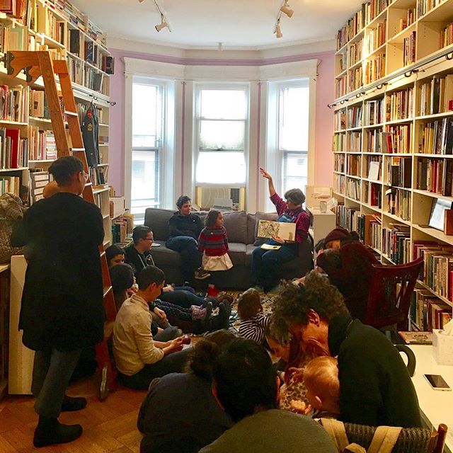 A group of adults and children are gathered on the floor of a room lined with bookshelves. A Lesbian sitting on a couch at the end of the room is reading a children's book to the group.