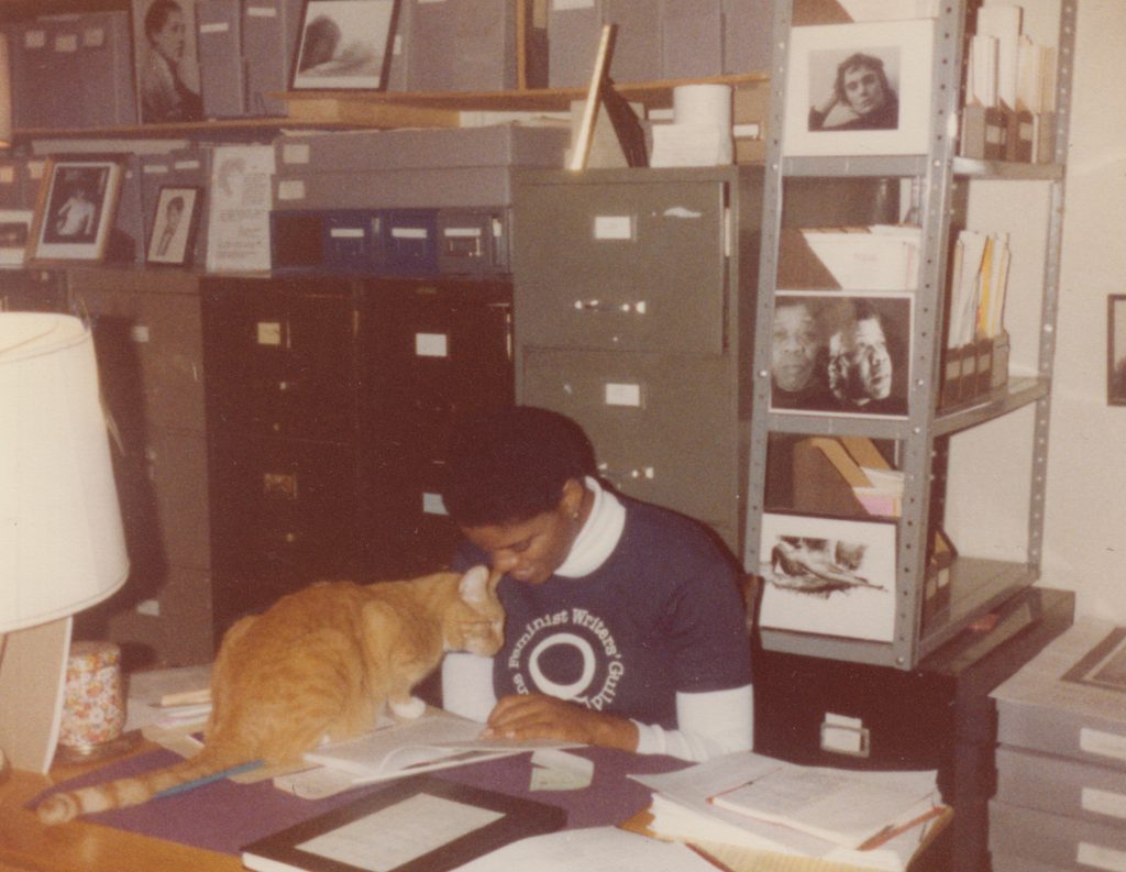 A Black Lesbian sitting at a table and an orange cat crouched on the table in front of her.