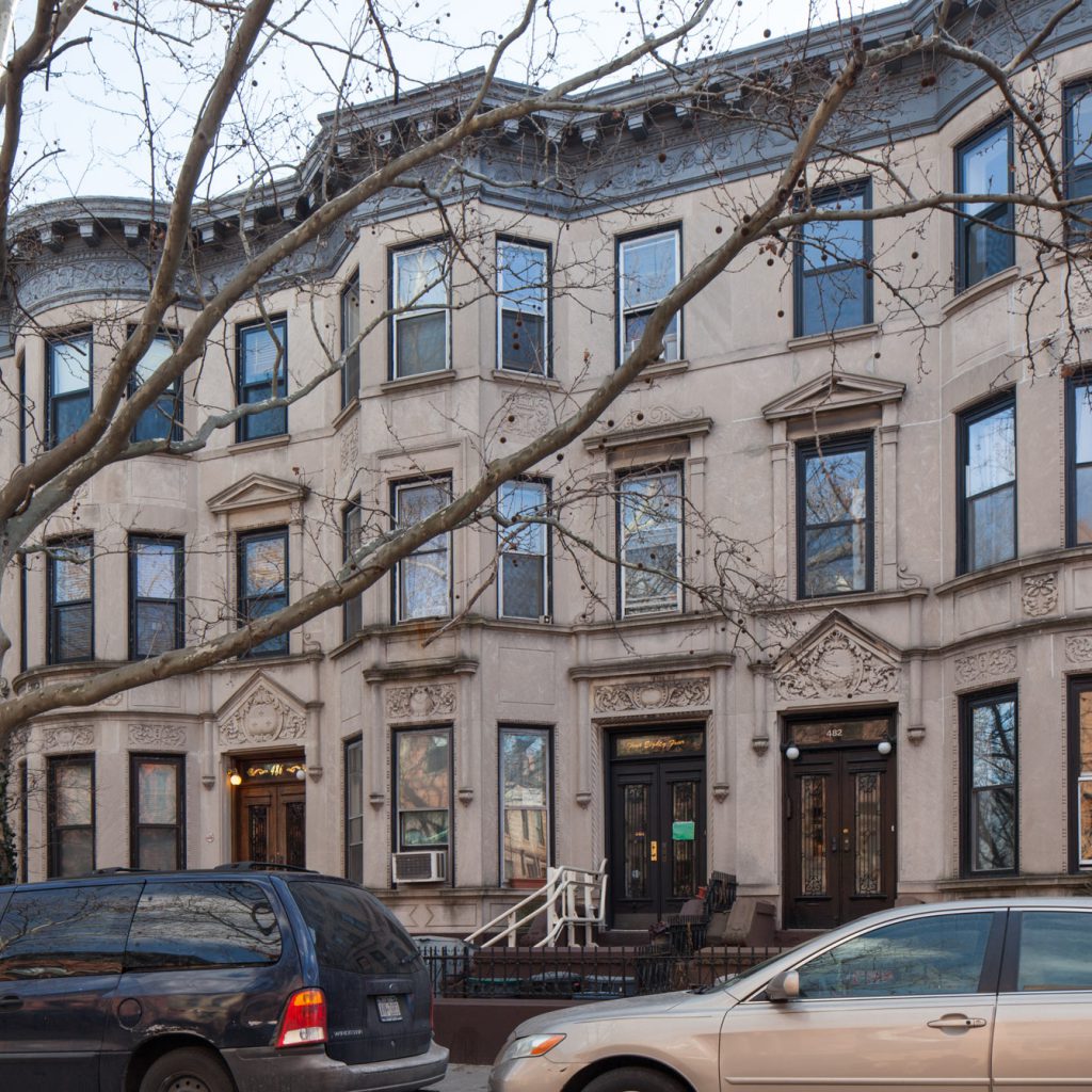 The facade of the Lesbian Herstory Archives is a smooth brown stone. The building has three stories, the first of which is decorated with ornate stonework above the doors. Taken in winter, this photo shows the barren limbs of a large tree in front of the Archives.