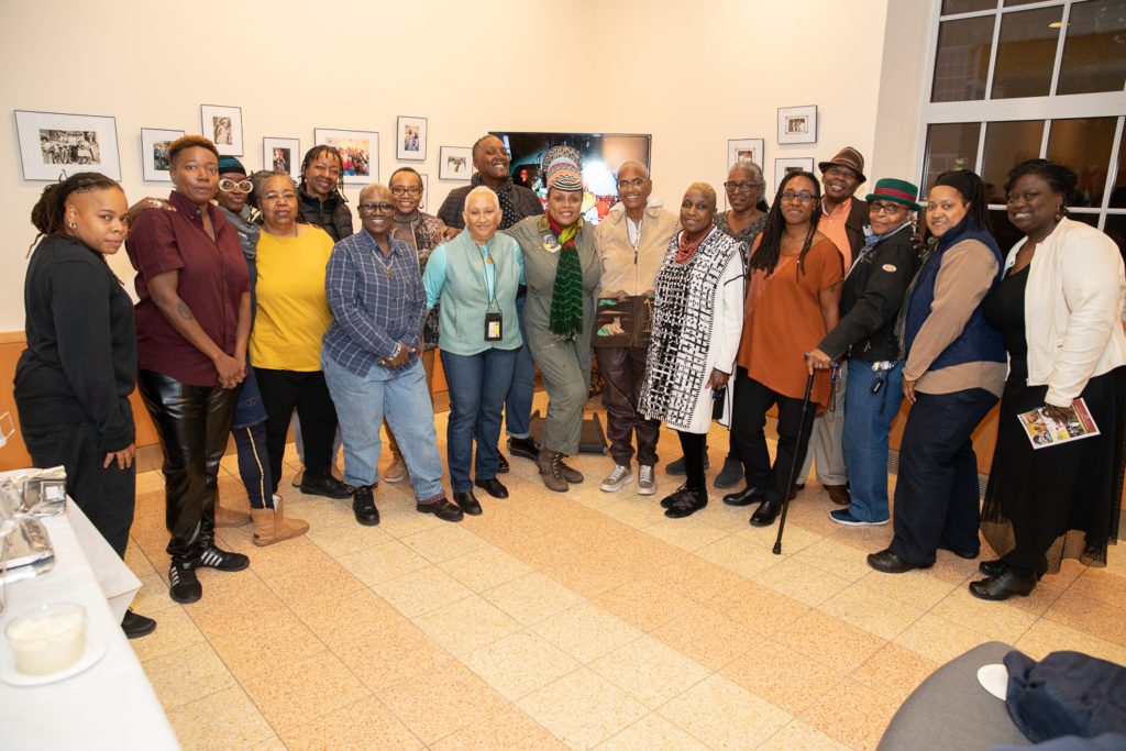 A group of eighteen Lesbians of Color stand together in a room, facing the camera and smiling. There are a number of framed photographs and other artwork on the walls behind them. 