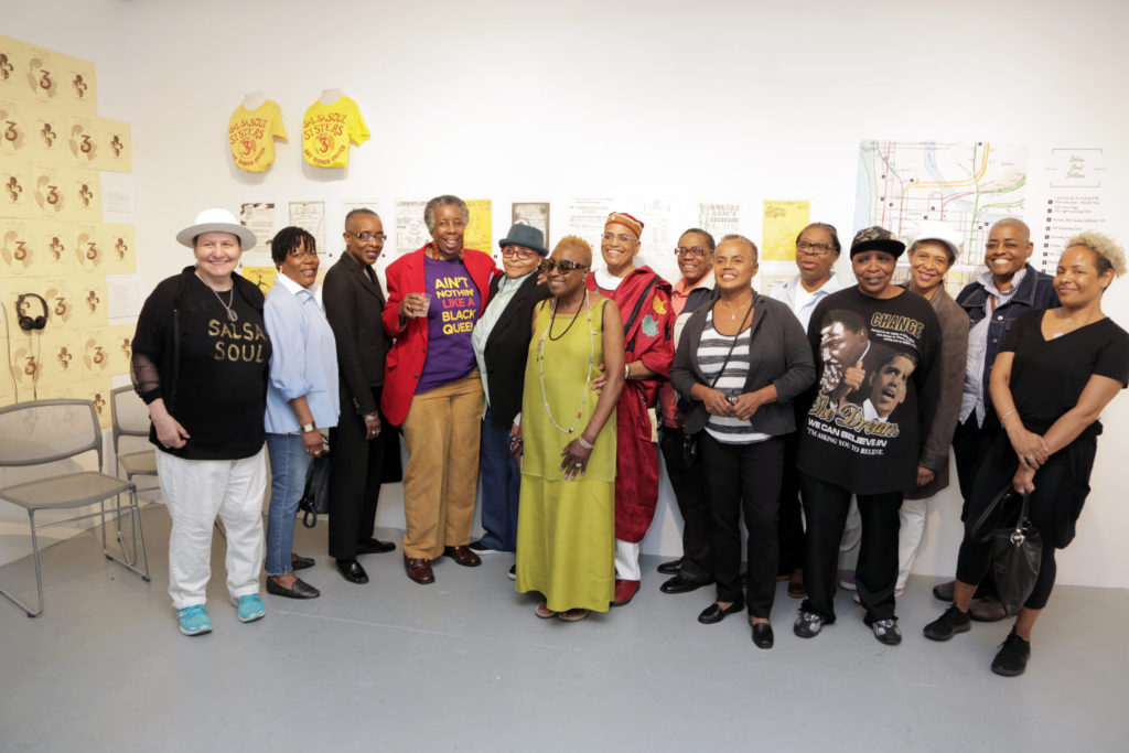 A group of fourteen Lesbians standing in front of a gallery wall.