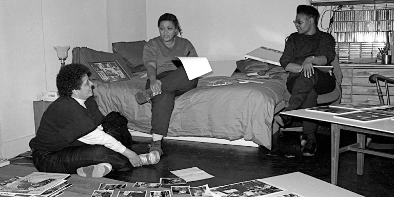 Three women sitting with papers open, in a working meeting, two on a bed one on the floor Working on the Keepin’ On exhibition. Pictured from left to right: Paula Grant, Jewelle Gomez, Georgia Brooks, 1991.