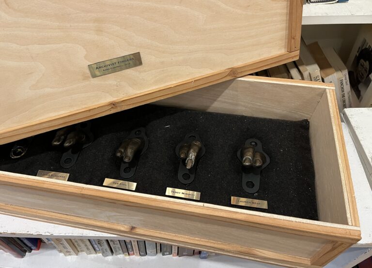 A wooden box sits on top of a low white shelf of books. On the front of the box is a small brass plague inscribed with "Archivist Fingers / Anna Campbell 2014." Inside the box are four bronze sculptures of the pointer and middle finger of four of the archivists who have served as Coordinators.