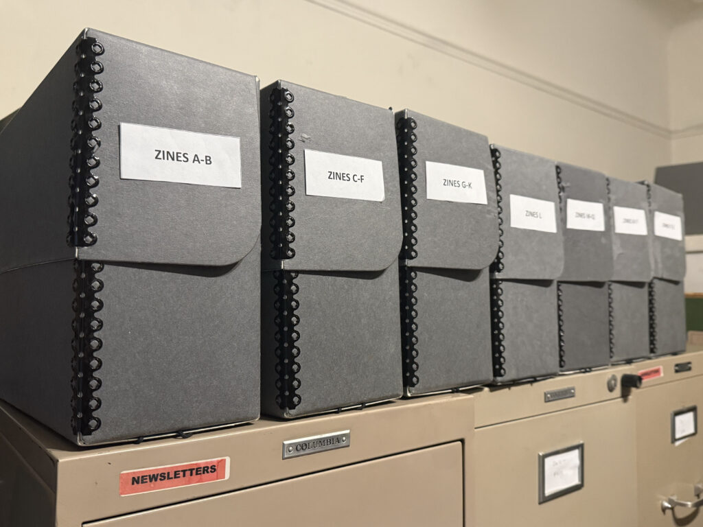 Five vertical archival boxes sit on top of filing cabinets. Each box has a label indicating that it holds zines and they are in alphabetical order. Example: "Zines A-B"